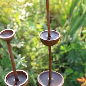 May include: A rusty metal sculpture with multiple round bowls, each filled with water, creating a cascading water feature. The sculpture is set against a backdrop of green foliage.
