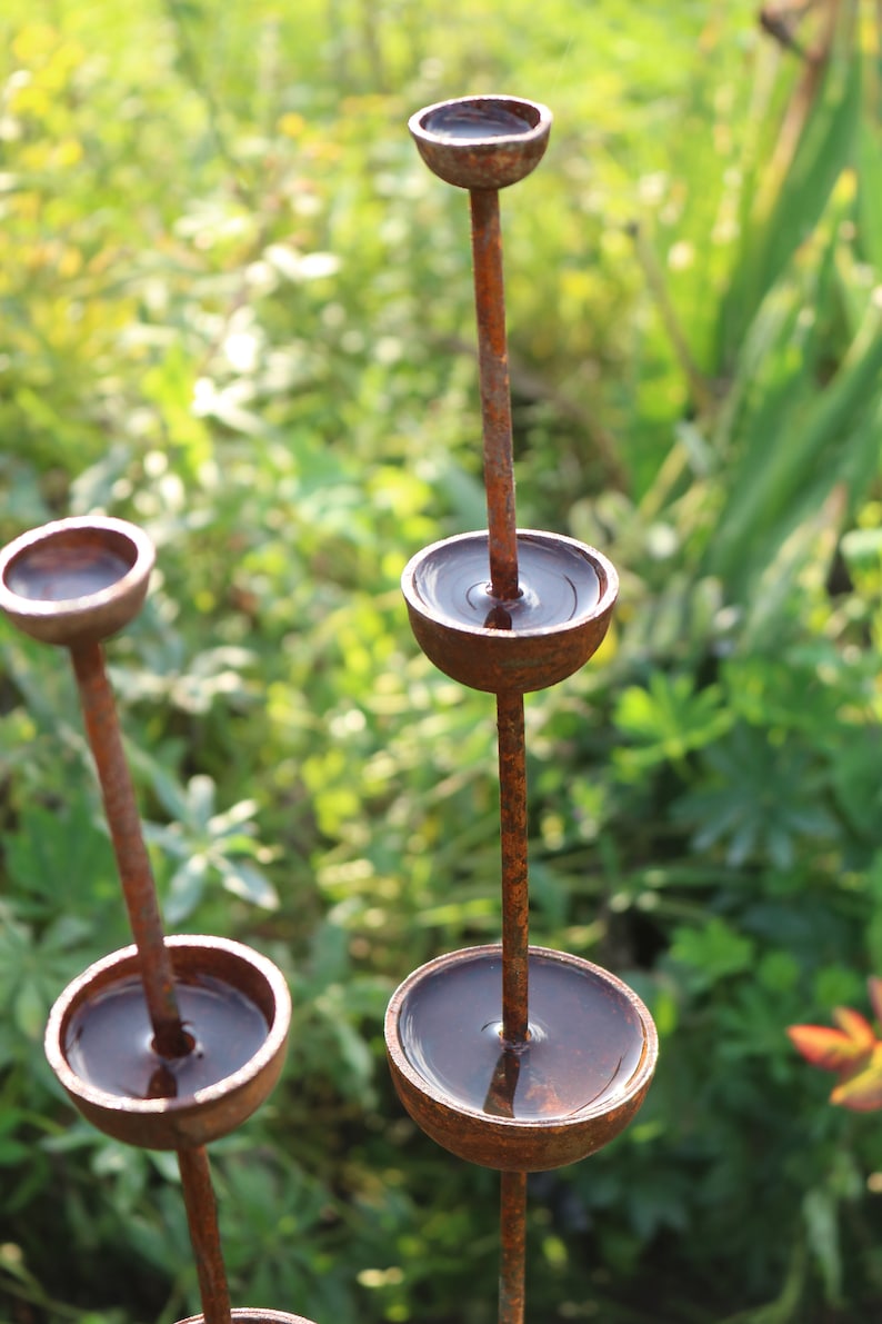 May include: A rusty metal sculpture with multiple round bowls, each filled with water, creating a cascading water feature. The sculpture is set against a backdrop of green foliage.