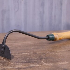 May include: A hand-held garden hoe featuring a semi-circular metal blade attached to a curved metal neck. The tool includes a light-coloured wooden handle with a dark green collar and a small circular hole at the end for hanging storage purposes.