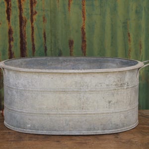 May include: An oval-shaped galvanised metal wash tub featuring a wide rim, two side handles, and horizontal ribbed detailing along the exterior walls. The basin has a weathered, matte silver-grey finish with visible signs of industrial use.
