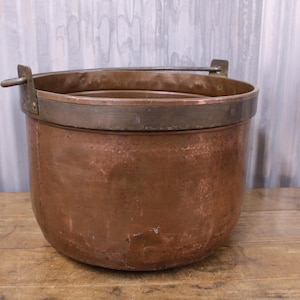 May include: A large, antique copper pot with a hammered finish and a metal band around the top. The pot has a handle on one side and is sitting on a wooden surface.