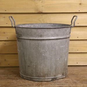 May include: A vintage galvanised metal bucket with two curved handles. The bucket is cylindrical with a slightly flared rim and a textured surface. The background is a wooden wall.