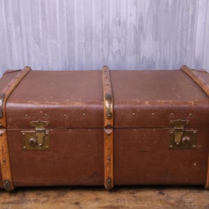 May include: A rectangular brown trunk featuring reinforced wooden slats, brass-toned metal corner protectors, and two central brass-toned locking latches. The exterior surface shows signs of wear, with visible rivets securing the wooden trim.