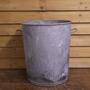 May include: A cylindrical galvanised metal bucket with a weathered, matte grey finish. The container features two small, angular metal handles attached to opposite sides near the top rim, providing a functional grip for lifting and carrying.