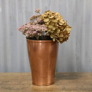 May include: A copper vase with a polished finish contains dried flowers. The vase has a tapered shape. The flowers include a large, dried hydrangea and smaller, pinkish-purple blooms. The vase is on a wooden surface.