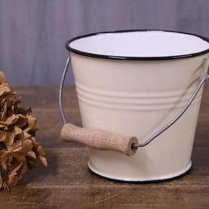 May include: A cream-coloured metal bucket with a black rim and a wire handle featuring a light brown wooden grip. The bucket has a smooth finish and a tapered cylindrical shape, accompanied by a cluster of dried brown hydrangea flowers.