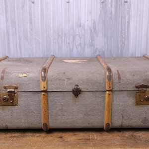 May include: A rectangular grey trunk featuring light brown wooden slats running vertically across the exterior. The trunk is secured with brass-coloured metal corner reinforcements, two side latches with keyholes, and a central metal locking clasp.