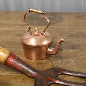 May include: A small copper kettle with a brass handle and spout, alongside a red and black garden fork with a wooden handle. The kettle is round, and the fork has three prongs.