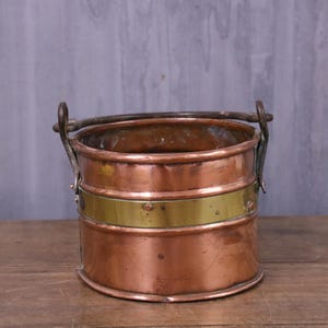 May include: A cylindrical copper bucket featuring a wide, horizontal brass band around the centre. The bucket has a dark metal swing handle attached to two side mounts and displays a warm, metallic finish with visible signs of age and use.
