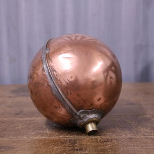 May include: A spherical copper float with a polished metallic surface, featuring a visible seam running around the centre and a small brass-coloured cylindrical attachment at the base. The copper shows slight signs of wear and natural oxidation.