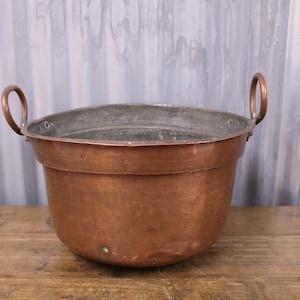 May include: A large copper pot with two handles. The pot is round and has a hammered finish. It is sitting on a wooden surface.