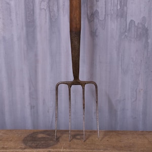 May include: A vintage garden fork featuring a long, weathered wooden handle attached to a dark, oxidised metal head with four slender, pointed tines. The metal neck shows signs of age and wear, connecting the handle to the pronged base.