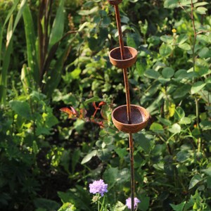 May include: A rusty metal garden sculpture with three round bowls stacked on a tall stem. The sculpture is surrounded by green foliage and purple flowers.