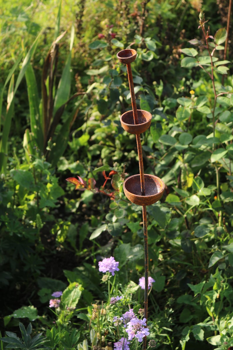 May include: A rusty metal garden sculpture with three round bowls stacked on a tall stem. The sculpture is surrounded by green foliage and purple flowers.