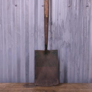 May include: A vintage shovel featuring a weathered, dark metal rectangular blade attached to a long, worn wooden handle. The metal surface shows signs of oxidation and age, while the wooden handle displays a natural, textured grain finish.