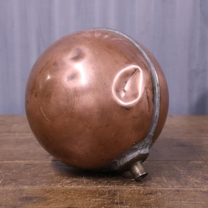 May include: A spherical copper float with a polished metallic surface featuring a visible indentation on the side. The object includes a silver-toned metal band encircling the circumference and a small cylindrical attachment at the base.