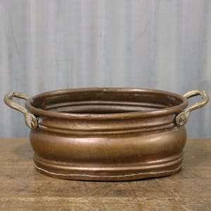 May include: A copper-coloured, round planter with two brass handles. The planter has a wide, shallow bowl shape with a slightly raised base and decorative horizontal ridges. The handles are curved and attached to the sides.