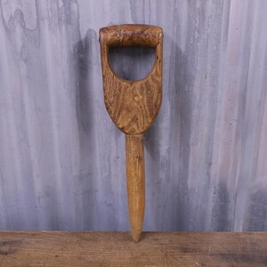 May include: A hand-carved wooden dibber featuring a natural light brown wood grain finish, a sturdy D-shaped handle at the top, and a tapered, pointed cylindrical shaft designed for creating planting holes in soil for seeds and bulbs.