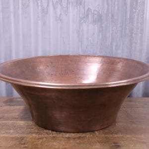 May include: A large, round, metallic copper basin with a wide, flat, rolled rim. The bowl features a smooth, polished surface with subtle variations in the warm, reddish-brown copper tone, tapering from a wide opening down to a flat base.