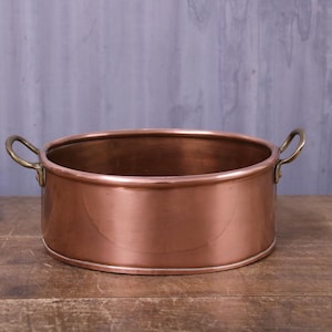 May include: A round copper pot with a polished metallic finish and two curved brass handles attached to opposite sides. The vessel features a smooth, wide rim and a flat base, showcasing the warm, reddish-brown tones of the polished copper material.
