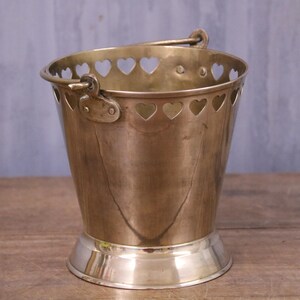 May include: A brass bucket with a tapered body and a wide, polished base. The upper rim features a series of cut-out heart shapes. A hinged metal handle is attached to the sides with circular rivets, allowing the handle to swing freely.