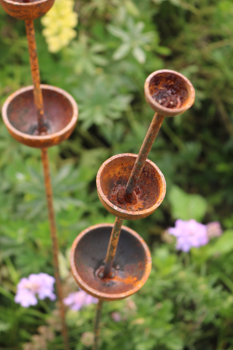 May include: A rusty metal garden sculpture with multiple round bowls stacked on top of each other. The sculpture is in a garden setting with green foliage and purple flowers.