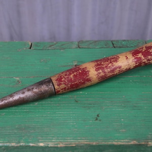 May include: A vintage hand tool featuring a pointed, weathered metal tip and a cylindrical wooden handle. The handle displays a worn, distressed finish with remnants of red paint over a natural light wood base, showing signs of age and use.