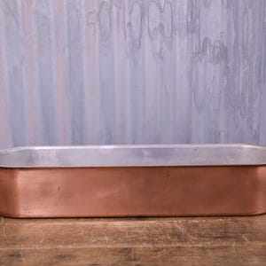 May include: An elongated oval roasting pan featuring a polished copper exterior and a silver-toned metallic interior. The vessel includes two curved brass-coloured handles attached to the sides with circular rivets, providing a sturdy grip.