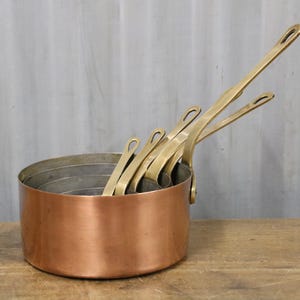 May include: A collection of copper saucepans with brass handles. The pans are stacked inside each other, with the handles pointing upwards. The copper has a warm, reddish-brown colour, and the brass handles have a golden hue. The pans are on a wooden surface.