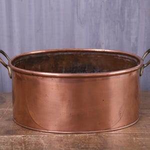 May include: A large, oval-shaped copper basin featuring a smooth, polished exterior surface and two sturdy, curved brass handles attached to opposite sides. The interior of the vessel shows a dark, weathered patina consistent with aged metal.