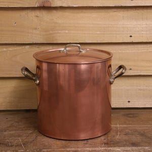 May include: A large, cylindrical copper stockpot with a polished metallic finish. The pot features two curved side handles made of a darker metal and a matching flat lid with a small, central handle on top for easy lifting and secure coverage.