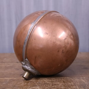May include: A spherical copper float with a polished metallic surface, featuring a central seam and a small, cylindrical brass-coloured attachment point at the base. The object has a smooth, rounded form with subtle variations in the copper tone.