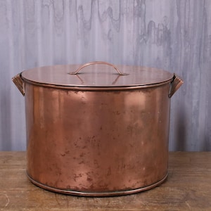 May include: A large, antique copper pot with a lid. The pot is cylindrical with side handles and a small handle on the lid. The copper has a warm, reddish-brown colour and shows signs of age and use, typical of vintage items.