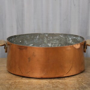 May include: A copper cooking pot with wooden handles. The pot has a wide, open design and a shiny, reflective surface. The interior of the pot has a dull, aged appearance. The pot is sitting on a wooden surface.