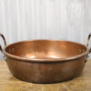 May include: A copper bowl with two arched handles. The bowl has a wide, shallow design and a polished surface. The copper has a warm, reddish-brown hue, and the handles are securely attached to the sides.