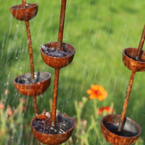 May include: A rusty metal garden fountain with multiple tiers of bowls, each catching water as it flows down. The fountain is surrounded by green grass and flowers.
