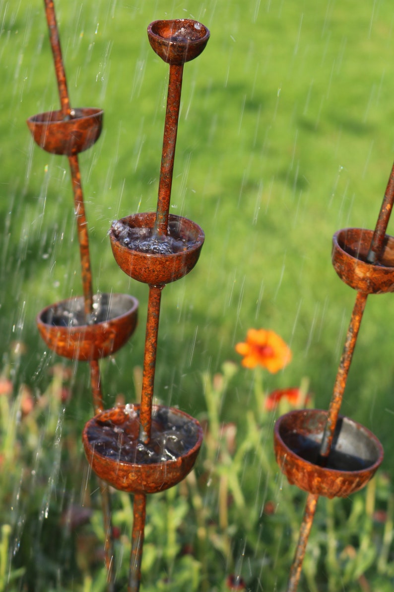 May include: A rusty metal garden fountain with multiple tiers of bowls, each catching water as it flows down. The fountain is surrounded by green grass and flowers.