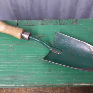 May include: A hand trowel featuring a dark green metal blade with visible rust along the curved edge, a green metal neck, and a light brown wooden handle with a smooth, worn finish. The tool is designed for gardening and digging tasks.