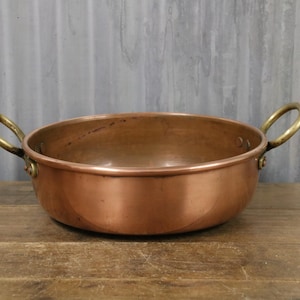 May include: A copper bowl with brass handles. The bowl is round with a wide, shallow design. The handles are attached to the sides of the bowl. The copper has a warm, reddish-brown colour, and the brass handles are a bright gold.