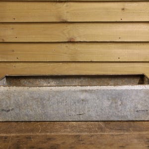May include: A rectangular, galvanised metal trough with a weathered, silver-grey finish. The container features straight, vertical sides and a flat base, showcasing a textured, industrial appearance with visible signs of age and oxidation.