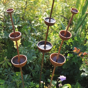 May include: A set of six rusty metal garden sculptures with rounded bowls on top of tall, thin stems. The bowls are filled with water and reflect the surrounding greenery.