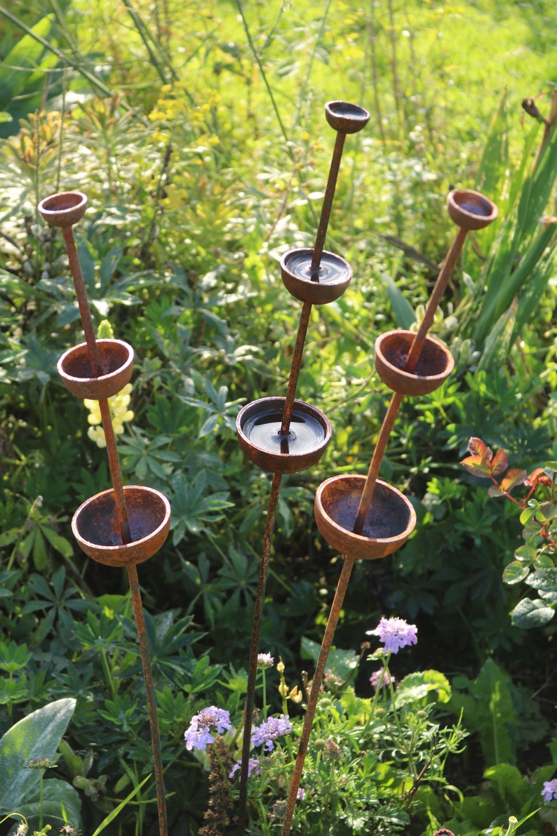 May include: A set of six rusty metal garden sculptures with rounded bowls on top of tall, thin stems. The bowls are filled with water and reflect the surrounding greenery.