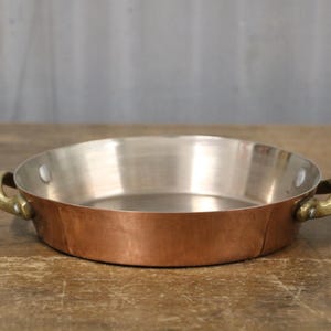 May include: A copper pan with a silver interior and brass handles. The round pan has a low profile, ideal for cooking or serving. The handles are curved and attached to the sides. The pan's diameter is approximately 25 cm.