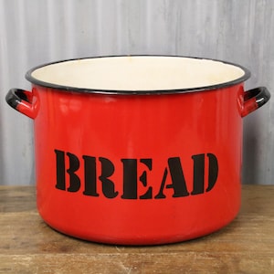 May include: A red enamel bread bin with black lettering spelling "BREAD". The bin has a black rim and handles. The interior is cream-coloured. The bread bin sits on a wooden surface.