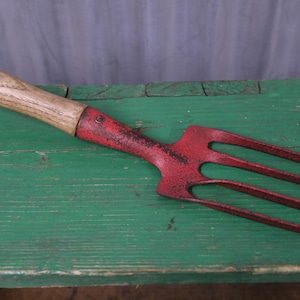 May include: A hand-held garden fork featuring a weathered red metal head with four tines and a light brown wooden handle. The metal surface shows signs of wear and texture, while the wooden handle displays a natural grain pattern and smooth finish.