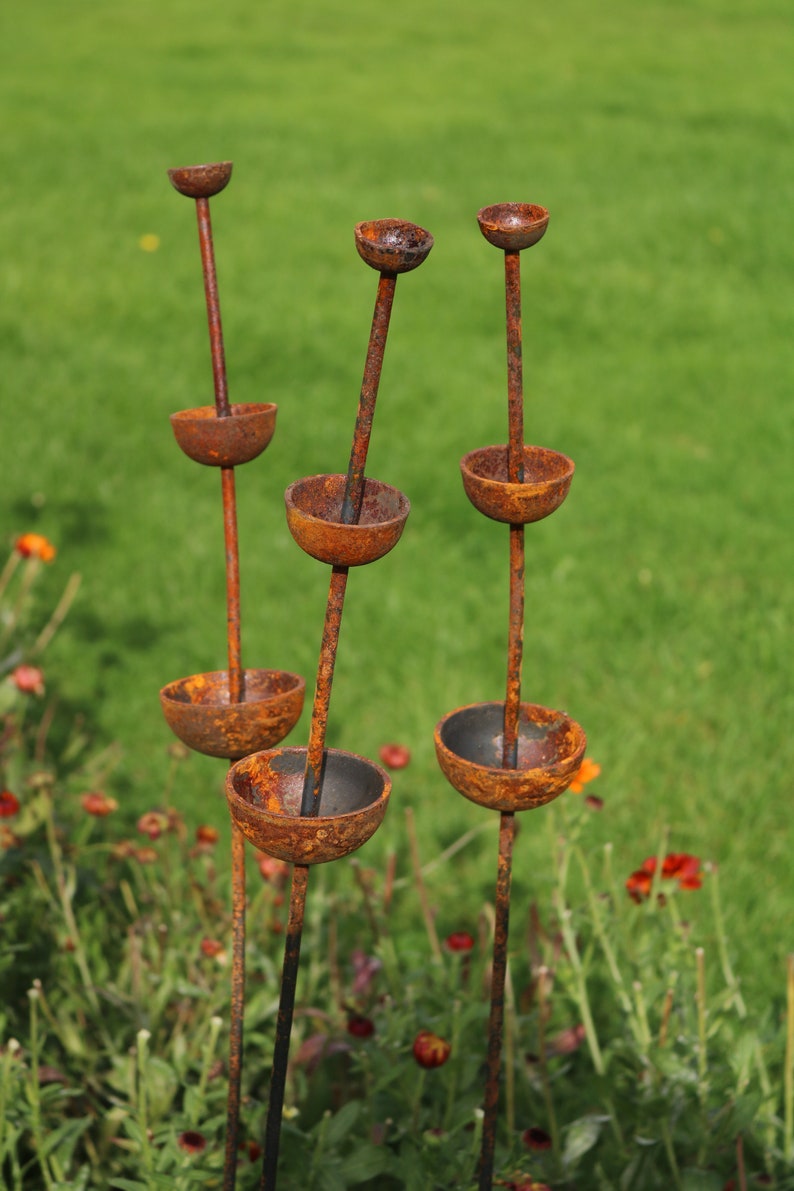 May include: Four rusty metal sculptures with rounded bowls stacked on top of each other. The sculptures are standing upright in a grassy field.