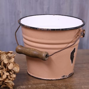 May include: A pale peach-coloured enamel bucket with a white interior and a black rim. The bucket features a metal handle with a cylindrical wooden grip. There is a visible black chip in the enamel coating on the lower side of the exterior surface.