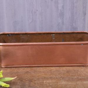 May include: A rectangular copper planter with rounded corners and a smooth metallic finish. The container features two small brass-toned metal ring handles attached to the sides, providing a simple and functional design for holding plants.