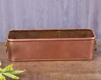 May include: A rectangular copper planter with rounded corners and a smooth metallic finish. The container features two small brass-toned metal ring handles attached to the sides, providing a simple and functional design for holding plants.