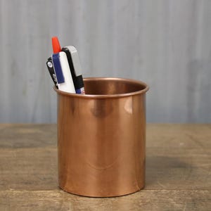 May include: A cylindrical copper pen holder with a rolled rim, containing pens and markers. The holder sits on a wooden surface against a grey background. The pens include red, black, and blue markers.